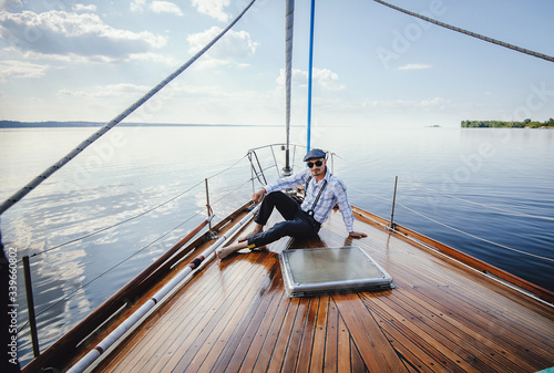 A successful businessman and yachtman resting on his yacht. Young man in sunglasses, shirt and cap sits on boat's nose