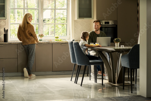 Smiling father working on laptop computer and talking to little cute daughter sitting at table in kitchen. Loving mother looking at them cooking at kitchen counter