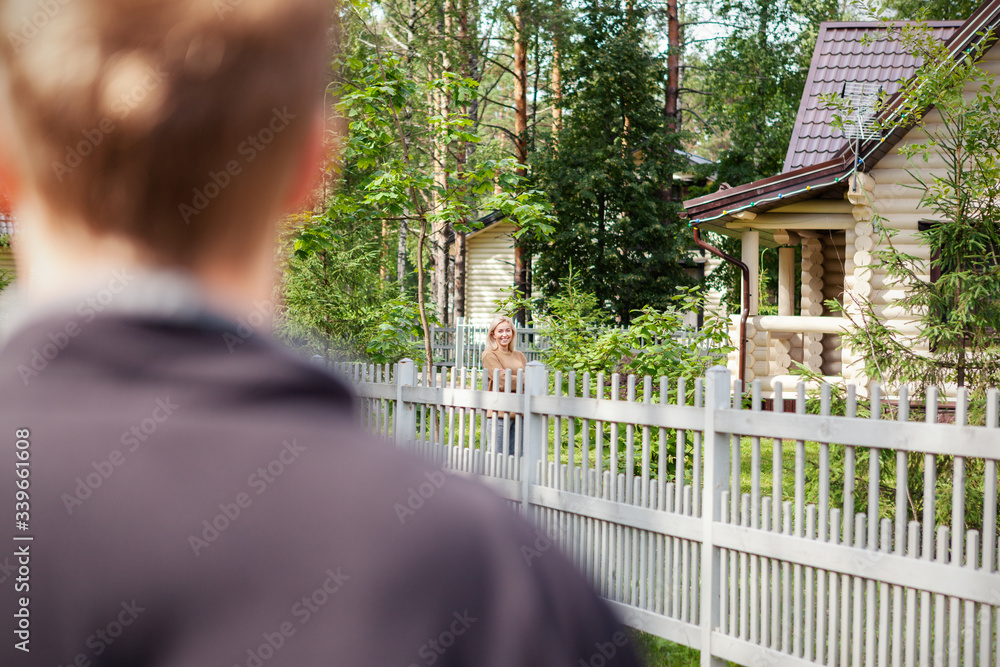 Over the shoulder view of defocused man walking in countryside and meeting female neighbor standing behind wooden fence, looking at him and smiling