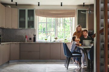 Middle aged man working on laptop computer in domestic kitchen surrounded by cute little daughter drinking juice and beautiful loving wife
