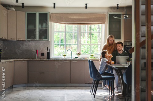 Middle aged man working on laptop computer in domestic kitchen surrounded by cute little daughter drinking juice and beautiful loving wife