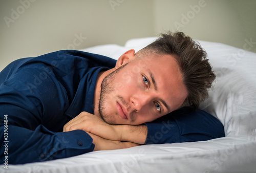 Sexy male model laying alone on bed in his bedroom, looking at camera with a seductive attitude,, wearing blue elegant shirt