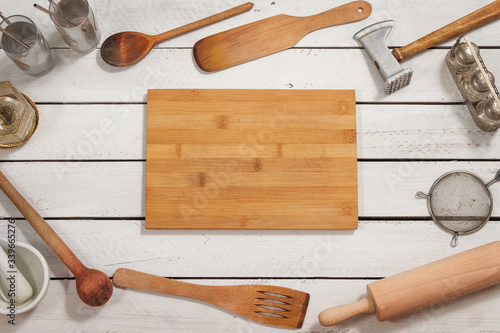 Drewniana deska kuchenna leży na białym stole. Na około leżą stare stylowe drewniane naczynia kuchenne tworzące tło.