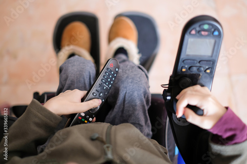 A disabled person sitting in her electric wheelchair chilling while holding the tv control watching television at home during social distancing