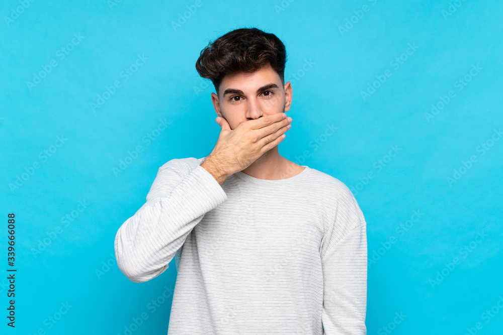 Young man over isolated blue background covering mouth with hands Young man over isolated blue background covering mouth with hands