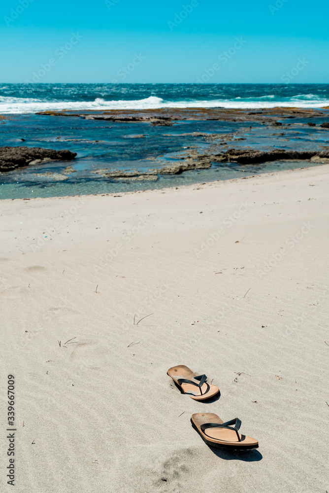 Flip flops on sandy beach at the seaside. Sea waves in the background. Empty beach. Vacation concept Flip flops on sandy beach at the seaside. Sea waves in the background. Empty beach. Vacation concept