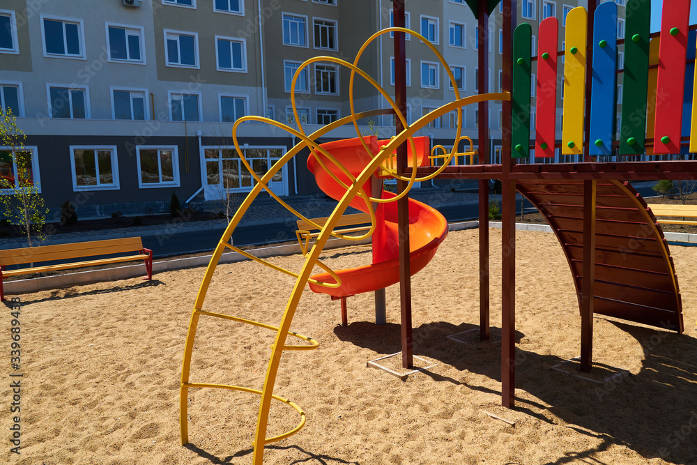 Urban residential infrastructure without people - children's playground next to a condominium. Swing, slide, stairs, multistory building. A place for children to play.