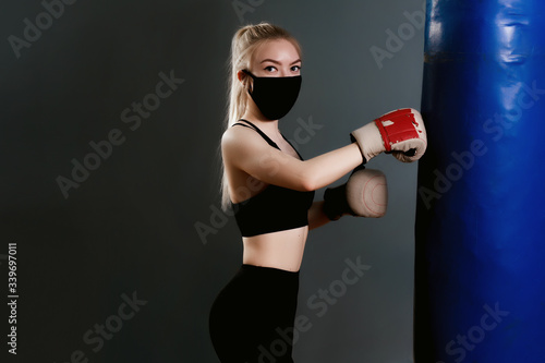 Young woman in a protective mask hits a punching bag. Protective masks against virus infection. Training during quarantine in the gym. Coronavirus (COVID-19) protection concept.