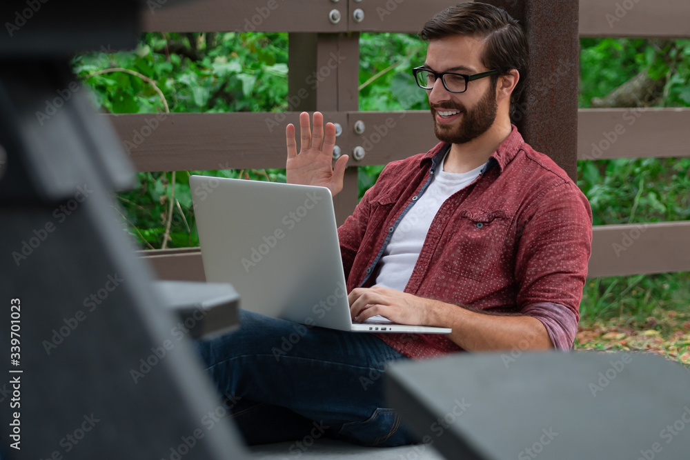 A young bearded hipster man with glasses sits against a pavilion pillar outside on a cloudy covered day near a walking path working remotely on his laptop waving trying to socially distance himself. A young bearded hipster man with glasses sits against a pavilion pillar outside on a cloudy covered day near a walking path working remotely on his laptop waving trying to socially distance himself.