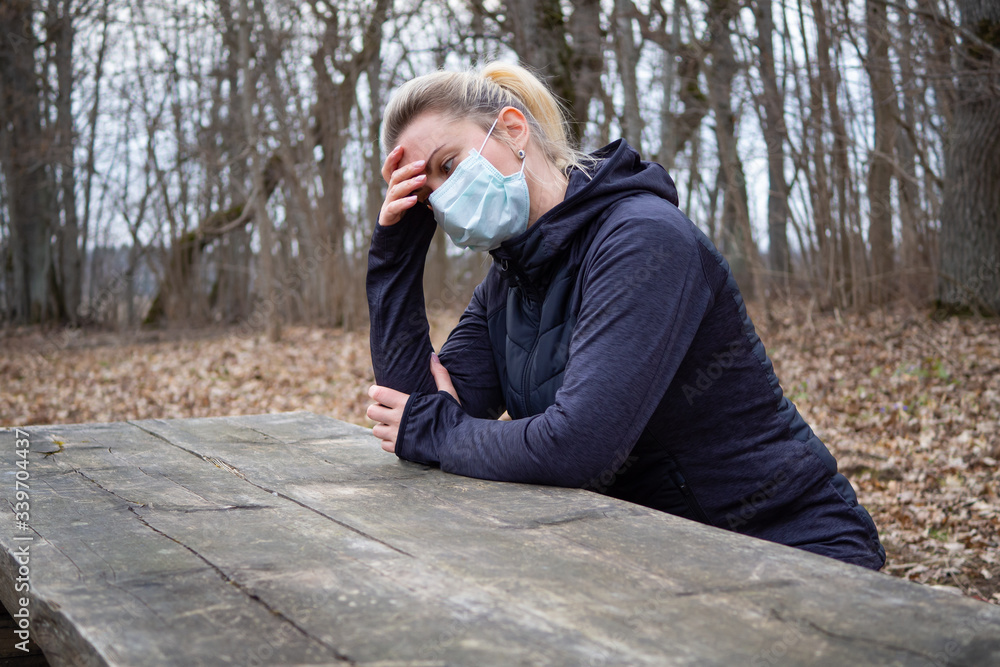 A young woman in a mask sits at a wooden table supporting head with hand. Girl resting in the park after training. Health prevention during isolation due coronavirus epidemic. Healthy lifestyle.