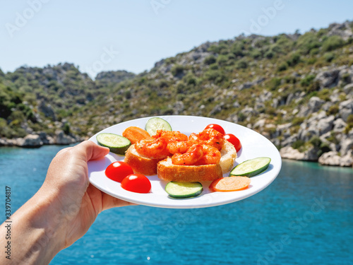 Mediterranean cuisine - freshly cooked shrimps on bread with vegetables. Hand with plate on beautiful sea lagoon background. Seafood with cucumbers and tomatoes.