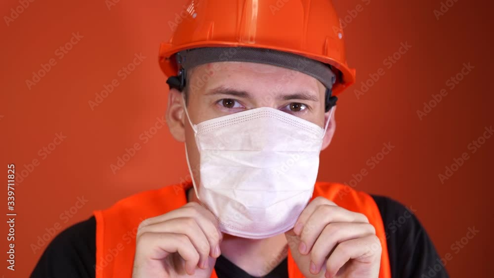 Male construction worker in overalls putting on medical mask on face on orange background. Male construction worker in overalls putting on medical mask on face on orange background.