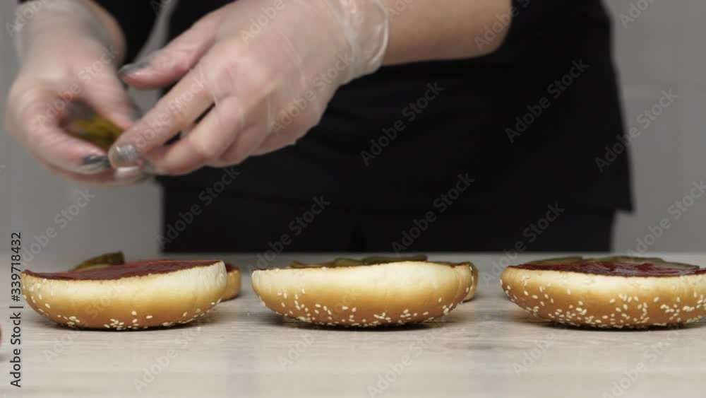 Top view of professional chef cooks a delicious burgers at fast food restaurant. Female chef in gloves puts pickles on three breads for burgers Top view of professional chef cooks a delicious burgers at fast food restaurant. Female chef in gloves puts pickles on three breads for burgers