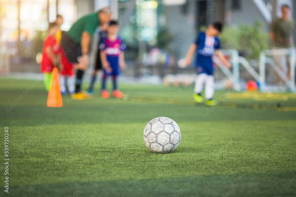 Old soccer ball on green artificial turf with blurry soccer team training. Blurry kid soccer player jogging between marker cones and control ball with soccer equipment in football academy. Old soccer ball on green artificial turf with blurry soccer team training. Blurry kid soccer player jogging between marker cones and control ball with soccer equipment in football academy.