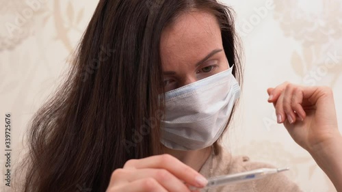 Beautiful Caucasian brunette ill girl in beige blouse wear blue protection medical face mask, take a thermometer out of her armpit. Russian woman look, check the high temperature at home close up.