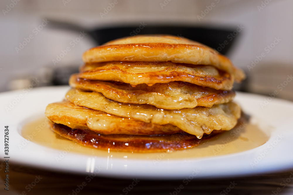 Fresh homemade classic pancakes with maple syrup in a white plate on a wooden board with a pan in the background. Fresh homemade classic pancakes with maple syrup in a white plate on a wooden board with a pan in the background.