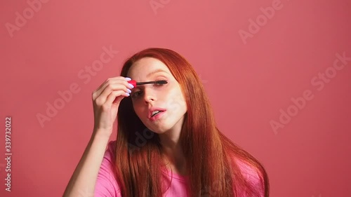 redhaired ginger woman in pink studio background holding mascara apply on eyeleashes looks at camera like at mirror photo