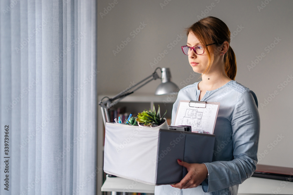 Young adult caucasian woman female girl standing by the window at the office losing her job holding personal items things in box being fired from work dismissed due to crisis recession quitting