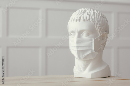 Plaster sculpture of Caesar's head stands on wooden table in medical mask. Protection against infections and coronavirus.