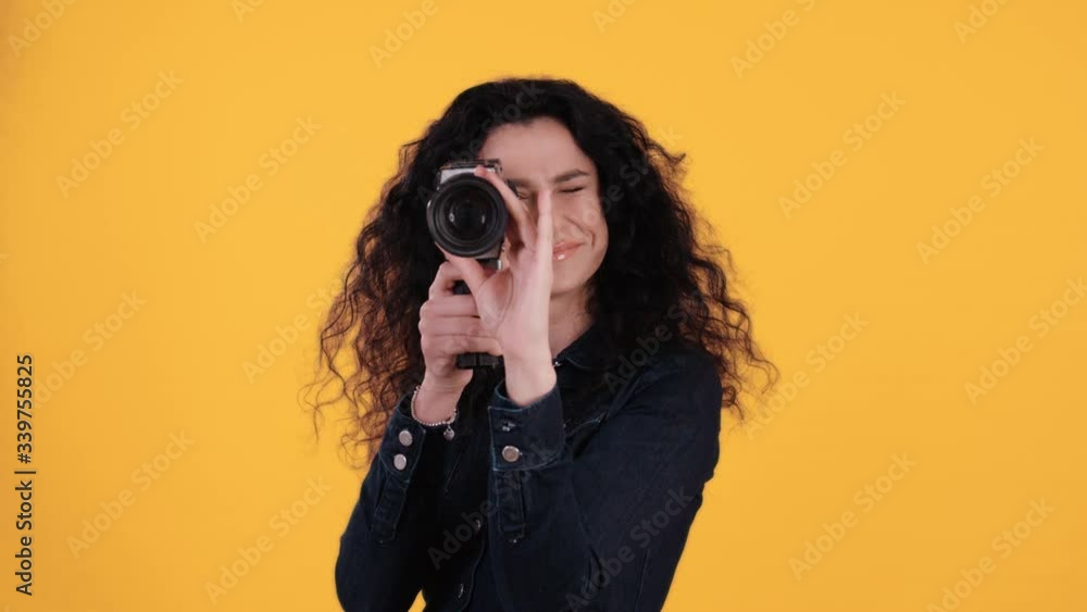 Portrait of beautiful young woman with black curly hair filing with oldschool retro 16mm cam towards the cam in front of yellow background shot in 4k Portrait of beautiful young woman with black curly hair filing with oldschool retro 16mm cam towards the cam in front of yellow background shot in 4k