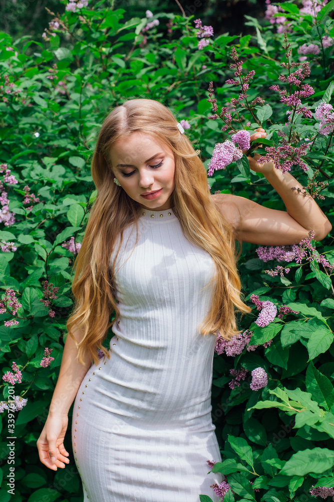 Portrait of a charming blond woman wearing beautiful white dress standing next to lilac bush. Portrait of a charming blond woman wearing beautiful white dress standing next to lilac bush.