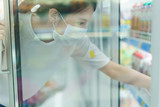 Woman with the surgical mask and gloves, looking for the drinks, in beverage cooler in grocery shop after coronavirus pandemic.Shot through glass