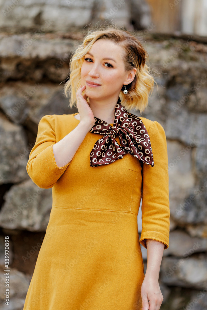 Fototapeta premium young attractive blonde in a mustard dress stands on the street and touches her face with her hands