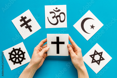 Choose religion concept. Hand with catholic cross near world religions symbols on blue background top view photo