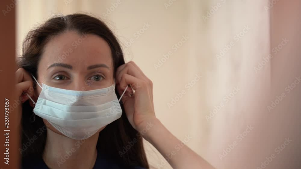Portrait of Young Beautiful Caucasian girl with dark brown hair with earrings looks at herself in the mirror, puts on blue medical face mask at home on white background close up. Woman going to work. Portrait of Young Beautiful Caucasian girl with dark brown hair with earrings looks at herself in the mirror, puts on blue medical face mask at home on white background close up. Woman going to work.