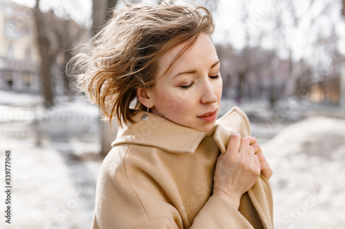 girl in a beige coat with blond loose hair wraps herself in a collar, the wind blows