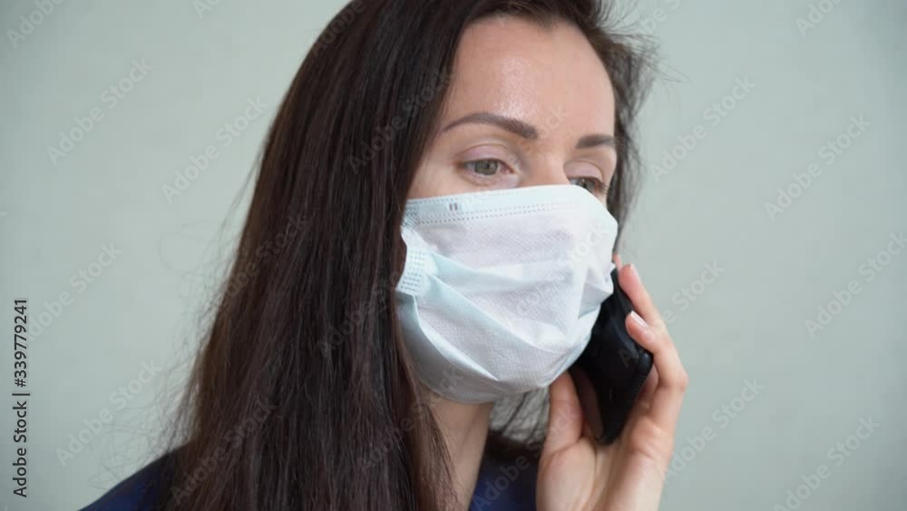 Beautiful Caucasian sick girl wear white protective face mask talking on mobile phone isolated on gray background close up. Young business woman with brown hair work remote. Protection by coronavirus. Beautiful Caucasian sick girl wear white protective face mask talking on mobile phone isolated on gray background close up. Young business woman with brown hair work remote. Protection by coronavirus.