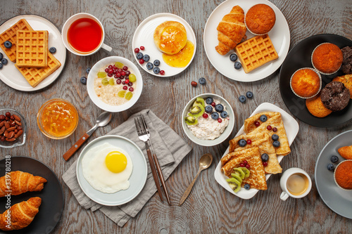 Continental breakfast captured from above (top view, flat lay). Coffee, tea, croissants, jam, egg, pancakes, maffins and oatmeal. Wooden background. Family breakfast table.