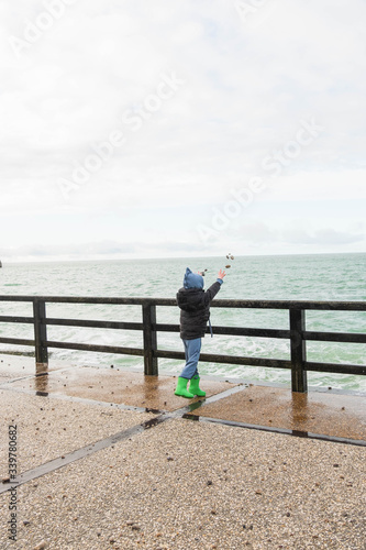 A small boy in a dragon costume throws pebbles into the ocean from the pier