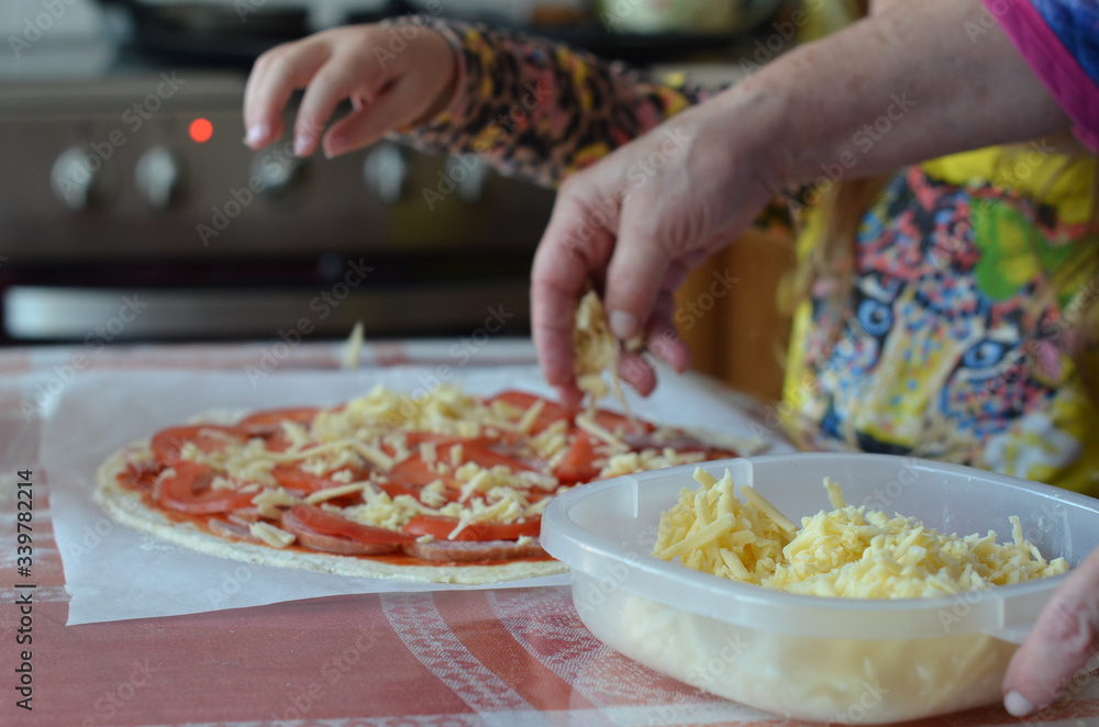 The hand of a woman and a child sprinkle grated cheese on an uncooked pizza. Joint cooking