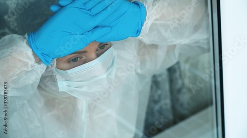 Sick alone isolated pensive girl in white protective suit, medical face mask, blue gloves stares longingly out through window at outside, leaning hands, head against glass. Coronavirus COVID-19 threat