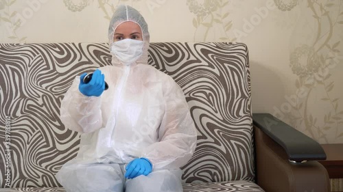 Alone woman in protective white suit in medical face mask in blue rubber gloves sits on couch, takes a remote control, switches channels, watching TV at home. Isolation during Covid-19 virus lock down