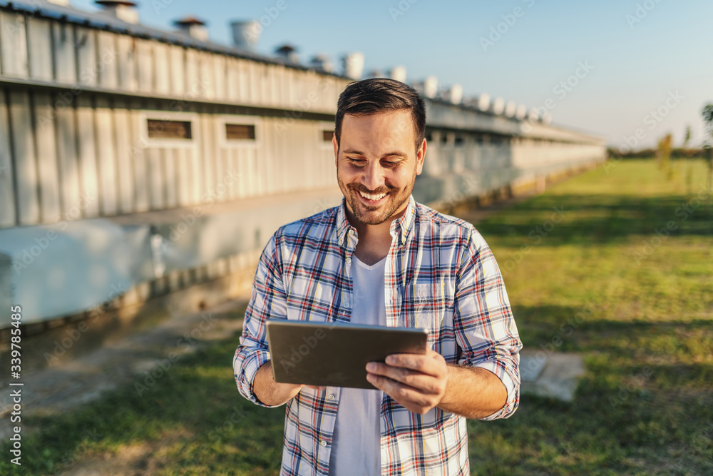 Fototapeta premium Cheerful caucasian farmer in plaid shirt standing outdoors and using tablet. In background are barns and orchard.