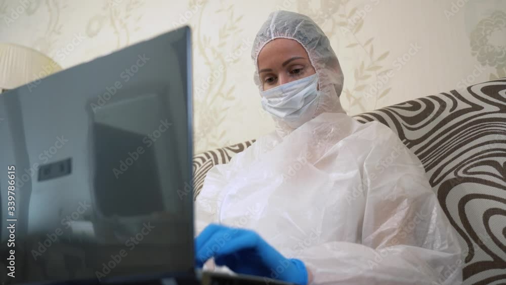 Young nurse wear white protective suit, medical face mask, blue gloves, using laptop. Female physician work remote at home in living room. Social distancing lockdown pandemic period by COVID-19 Young nurse wear white protective suit, medical face mask, blue gloves, using laptop. Female physician work remote at home in living room. Social distancing lockdown pandemic period by COVID-19