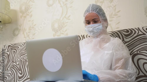 Sick young infected pretty girl isolated from society at home. Alone Ill woman in white protective suit, face mask, blue gloves using laptop, writing, surfing net. Remote work in quarantine isolation. photo