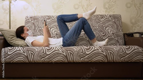 Attractive Caucasian brunette woman in protective medical face mask, relaxes on a lounge, using mobile phone, stretches, hold in hand remote control, switch channels, watch TV. Lazy girl in isolation.