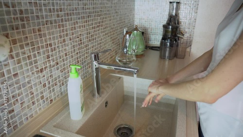 Close up of young brunette woman in protective medical face mask clean hands in pure water from tap or faucet, using hand sanitizer gel, perform morning cleanup routine in kitchen at home. Prevention photo