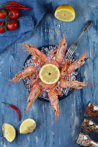 Plate of king prawns in ice with lemon and seasonings on a blue background. Tomatoes, hot peppers, lemon and dried seasonings in the mills around a blue plate with juicy shrimps. Seafood top view.