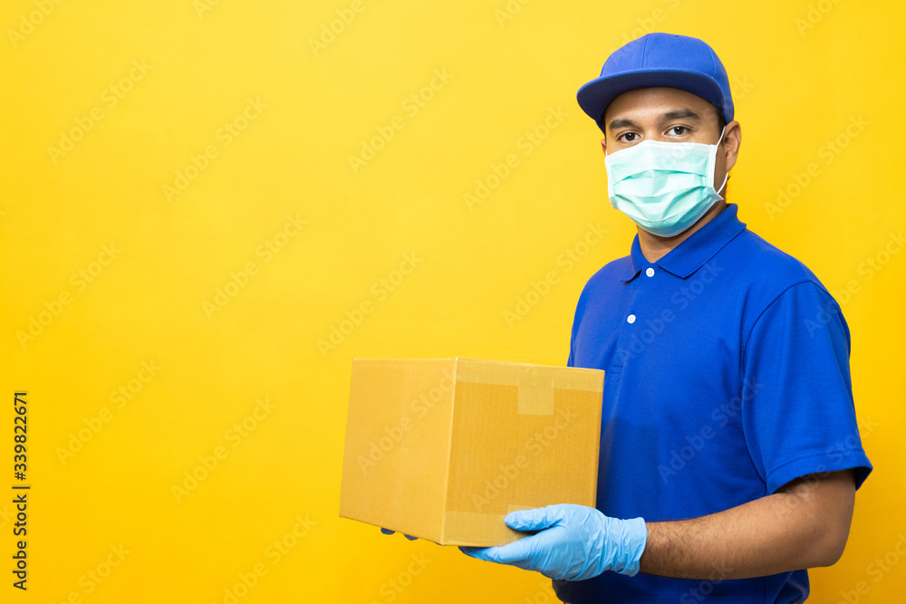 Delivery man blue uniform wearing rubber gloves and mask holding parcel cardboard box on yellow background. Delivery man blue uniform wearing rubber gloves and mask holding parcel cardboard box on yellow background.