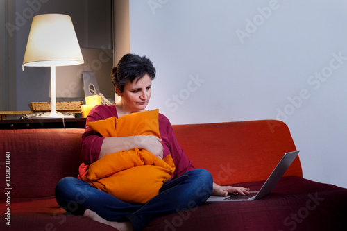 Donna Bianca con i capelli scuri, vestita casual, abbracci il cuscino seduta nel divano mentre guarda una serie tv dal portatile photo