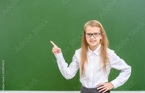 Smart girl wearing eyeglasses points away on empty green chalkboard. Empty space for text