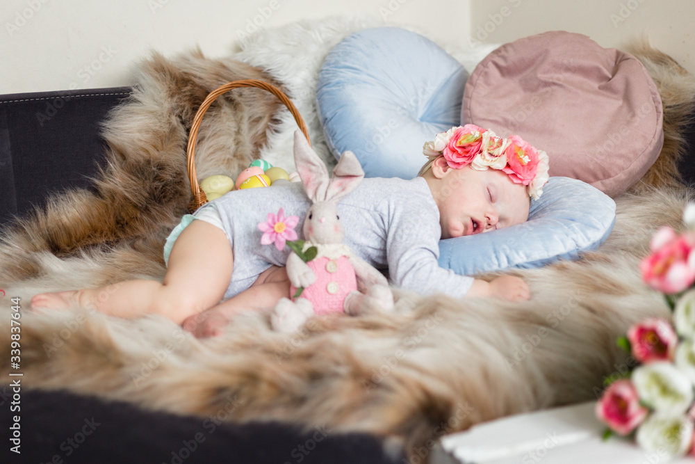 Little baby girl in flower crown, sleeping on a sofa on blue pillow and artificial fur, surrounded by easter decorations - bunny, tulips and basket with eggs