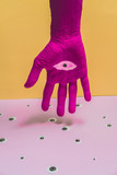 Painted in fuchsia pink hand with a drawn eye in the middle of the palm on a light yellow and purple or lilac background among many eyes. Abstract modern minimal pop art.