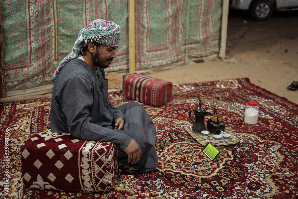 Bedouin man wearing traditional clothes watches a footbal match in a smartphone on a carpet in the Saudi desert