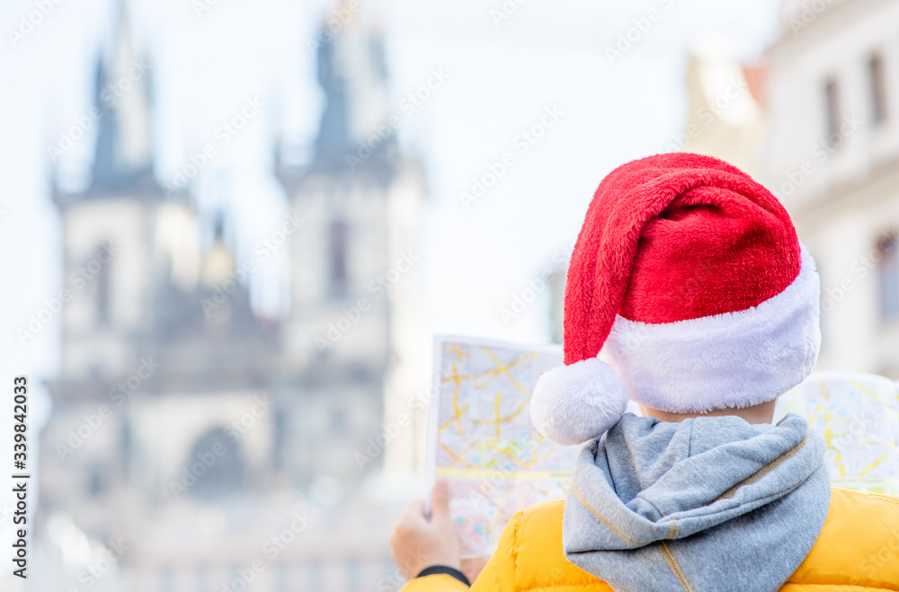 Naklejka premium Young boy wearing red santa's hat stands on the old town square in Prague and holds map. Empty space for text