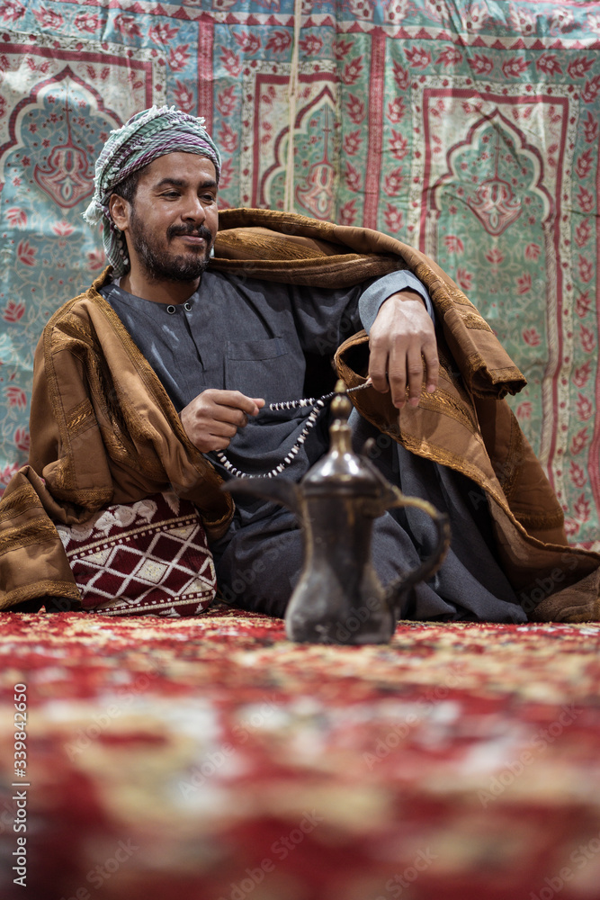 Bedouin man wearing traditional clothes praying with a tasbih while drinking tea on a carpet in the Saudi desert